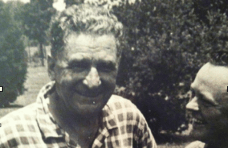 Black and white photo of a smiling man in a checkered shirt outdoors.